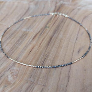 May include: Delicate beaded necklace featuring a mix of gold and dark gray beads. The necklace has a gold-toned chain and clasp, and is displayed on a wooden surface. This piece of jewelry is suitable for everyday wear or special occasions.
