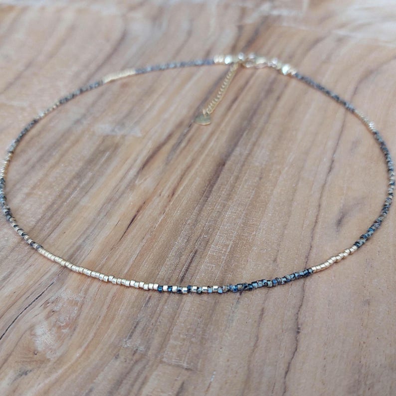 May include: Delicate beaded necklace featuring a mix of gold and dark gray beads. The necklace has a gold-toned chain and clasp, and is displayed on a wooden surface. This piece of jewelry is suitable for everyday wear or special occasions.
