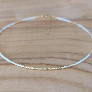 May include: Delicate beaded choker necklace. The necklace features small white and gold beads. The gold beads are concentrated in the center, with white beads on either side. The necklace has a gold-colored chain and clasp.