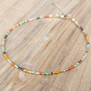 May include: A delicate beaded anklet featuring a mix of colorful beads in shades of orange, brown, yellow, green, and silver. The anklet is made with small, square beads and has a gold-colored clasp. The anklet is displayed on a wooden surface.