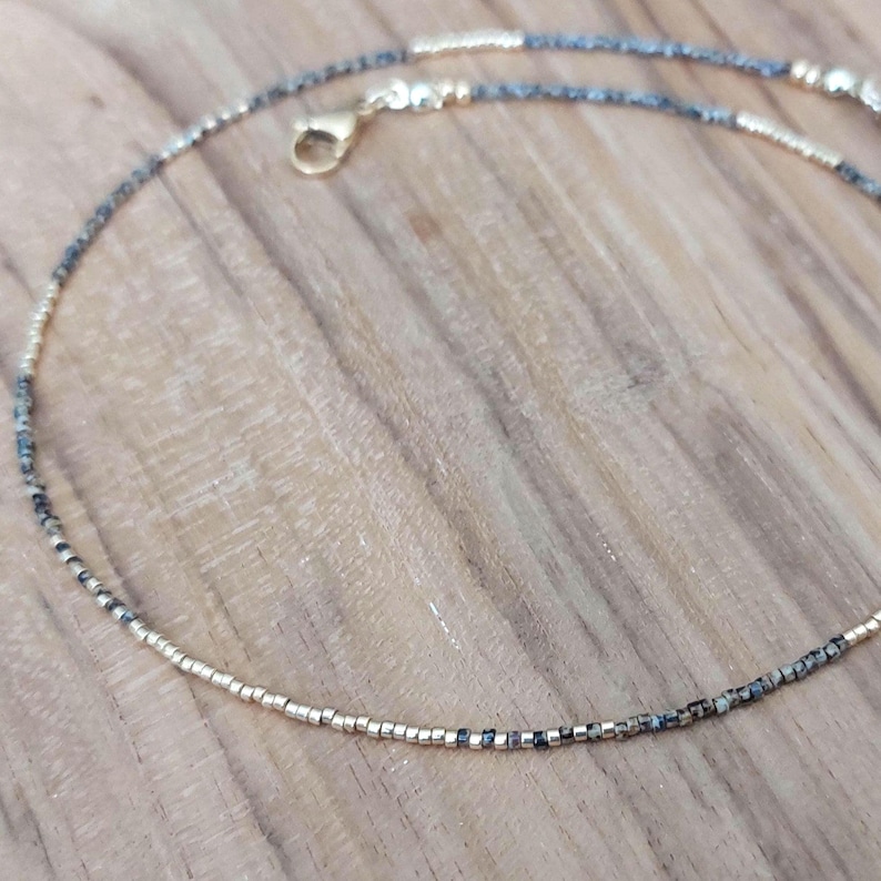 May include: Delicate beaded necklace featuring a mix of gold and dark gray beads. The necklace has a gold-colored clasp and is displayed on a wooden surface. A simple and elegant jewelry piece.