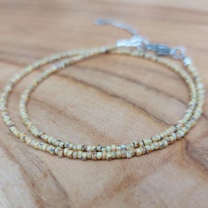 Op de afbeelding: Een delicate kralenketting met een zilveren sluiting. De ketting is gemaakt van kleine, lichtgekleurde kralen met subtiele kleurvariaties, waardoor een textuur ontstaat. De kralen zijn geregen tot een dubbele streng.