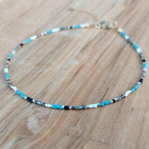 May include: Delicate beaded necklace featuring a pattern of turquoise, black, white, and gold beads. The necklace is strung on a gold-colored chain and rests on a wooden surface. A stylish accessory for everyday wear.