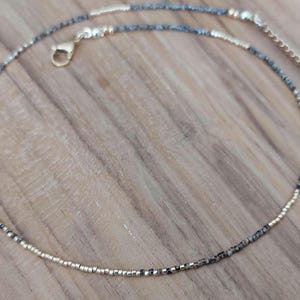 May include: Delicate beaded anklet featuring a mix of gold and speckled gray beads. The anklet has a gold-colored clasp and adjustable chain. The beads are small and evenly spaced, creating a refined and elegant look. This jewelry piece is perfect for everyday wear.
