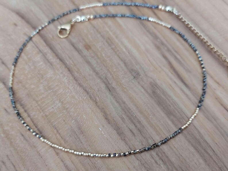 May include: Delicate beaded anklet featuring a mix of gold and speckled gray beads. The anklet has a gold-colored clasp and adjustable chain. The beads are small and evenly spaced, creating a refined and elegant look. This jewelry piece is perfect for everyday wear.