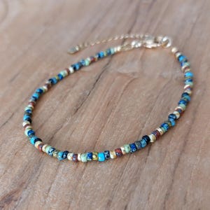 Op de afbeelding: Een delicate armband met een doorlopende streng van kleine, veelkleurige glaskralen in tinten blauw, groen, rood en crème, afgewisseld met kleine goudkleurige metalen tussenstukjes en een goudkleurige metalen karabijnsluiting.