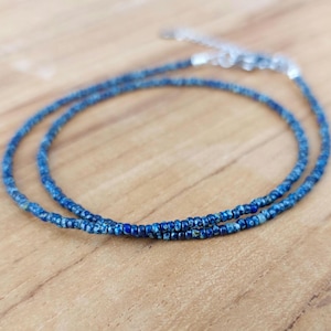 May include: A fine necklace composed of tiny, round, blue beads. The beads are strung to create a double strand, with a silver-coloured clasp. The necklace is presented on a light brown wooden surface.