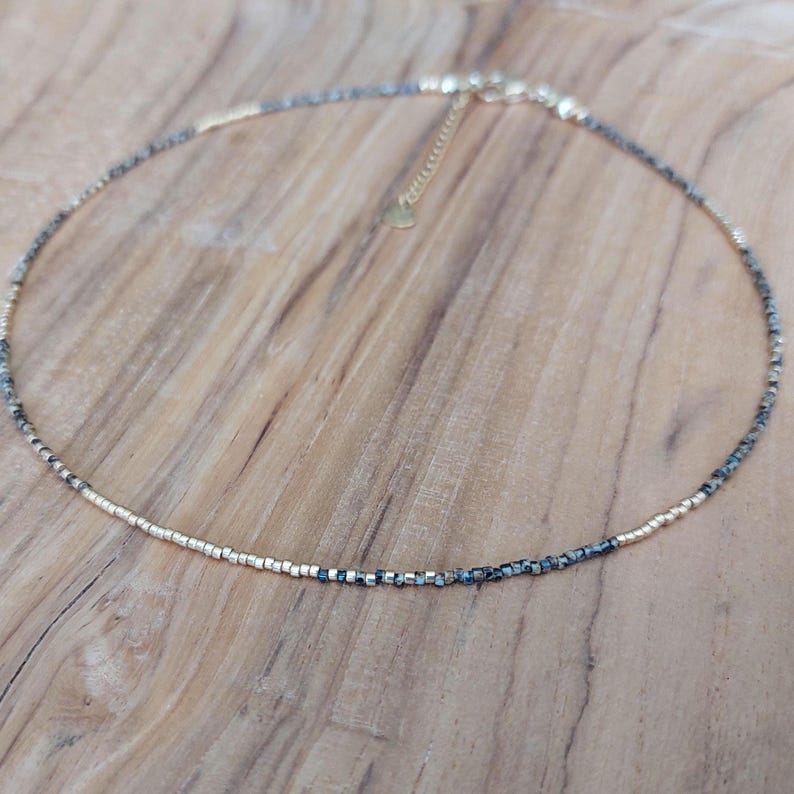 May include: Delicate gold-toned necklace with a beaded design. The necklace features small, dark gray and gold beads, creating a subtle contrast. The necklace is displayed on a wooden surface.