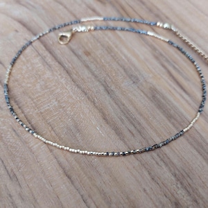 May include: Delicate gold and beaded necklace with a small clasp. The necklace features a mix of gold and dark gray beads, creating a subtle contrast. The necklace is designed to be worn around the neck.