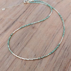 May include: Delicate necklace featuring small, turquoise-colored beads interspersed with silver-toned beads. The necklace has a simple design with a delicate chain and clasp, perfect for layering or wearing alone. The necklace is displayed on a wooden surface.