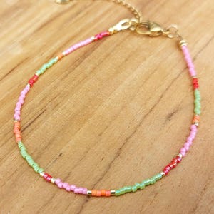 Op de afbeelding: Een delicate armband gemaakt van kleine, veelkleurige kraaltjes in tinten roze, lichtgroen en oranje, geaccentueerd met kleine goudkleurige metalen tussenstukjes. De armband is voorzien van een goudkleurige karabijnsluiting en een verstelbare ketting.