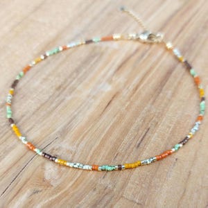 Op de afbeelding: Een enkelbandje met kralen en een goudkleurige sluiting en verlengketting. Het enkelbandje heeft kleine, kleurrijke kralen in tinten oranje, geel, bruin, groen en wit. Het enkelbandje wordt gepresenteerd op een houten oppervlak.