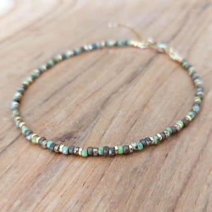 Può includere: Delicato braccialetto di perline con piccole perle rotonde nei toni del verde, blu e grigio, intervallate da distanziatori color oro. Il braccialetto è su una superficie di legno.