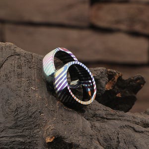 Könnte beinhalten: Zwei Ringe mit einem gestreiften Muster in Lila, Blau, Rosa und Gelb. Die Ringe bestehen aus einem glänzenden, metallischen Material und liegen auf einer dunklen, strukturierten Oberfläche.
