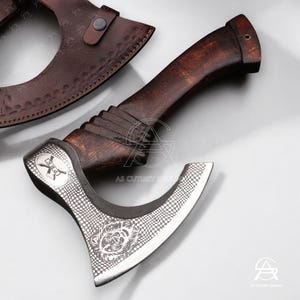 May include: A handcrafted axe with a dark wooden handle and a silver-colored head. The head features a detailed pattern and a decorative emblem. A brown leather sheath is visible in the background. The axe is designed for utility and display.