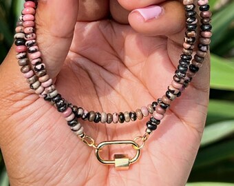 May include: A beaded necklace with a gold clasp. The necklace features small, round beads in shades of pink, brown, and black. The clasp is a gold-colored rectangle. The necklace is held in a hand with pink nail polish.