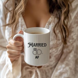 Puede incluir: Taza de café de cerámica blanca con texto negro que dice "MARRIED AF" y un gráfico de dos anillos de boda con un diamante en uno de los anillos.