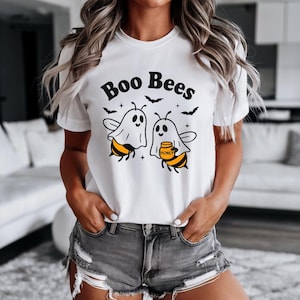 May include: White t-shirt featuring the phrase "Boo Bees" above a graphic of two bees in ghost costumes. One bee holds a honey pot. The design also includes black bats and stars.