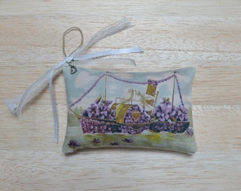 May include: A small white fabric sachet with a lavender and floral print of a boat with two birds on top. The sachet has a white lace ribbon attached to it.