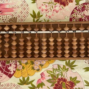 May include: A wooden abacus with brown beads and a white label with numbers. The abacus is on a patterned fabric background.