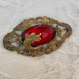 May include: An oval-shaped brass brooch featuring an ornate, filigree-style metal frame with a central, faceted, deep red gemstone. A sculpted brass dragon figure is positioned over the lower left portion of the vibrant red stone.