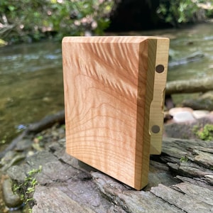 May include: A rectangular wooden block with a light brown, wavy grain pattern on its front face. The side profile reveals a lighter-coloured wood edge featuring two small, circular, dark-coloured magnetic inserts embedded into the surface.