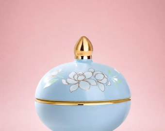 May include: A light blue ceramic jar with a gold-colored lid and trim. The jar is decorated with white and gold floral designs and the word "Jasmine" in black script. The jar is set against a pink background.