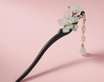 May include: A black wooden hair stick with a floral design. The hair accessory features white and pale green flowers, a gold chain, and dangling pearls and a teardrop-shaped stone. The background is a solid pink.