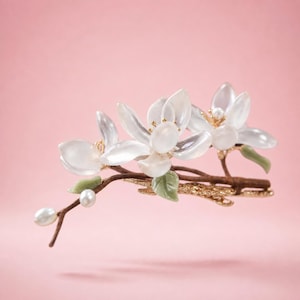 以下が含まれることがあります： 3つの白く半透明な花のつぼみ、金色のアクセント、緑の葉、イミテーションパールをあしらったフローラルヘアアクセサリー。 花は、金色のディテールが施された茶色の枝に取り付けられ、ピンク色の背景に配置されています。