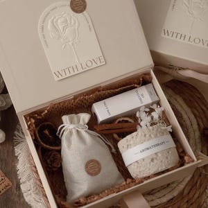 Op de afbeelding: Crèmekleurige geschenkdoos met een klein zakje met trekkoord, een zeepblok met het woord "AROMATHERAPY", een flesje essentiële olie en gedroogde sinaasappelschijfjes. De doos heeft een kaart met "WITH LOVE".