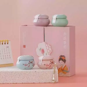 May include: A collection of decorative jars in pastel colors, including pink, blue, and green, with lids. The jars are adorned with floral and bird designs. A pink box with Chinese characters and a calendar are also present.