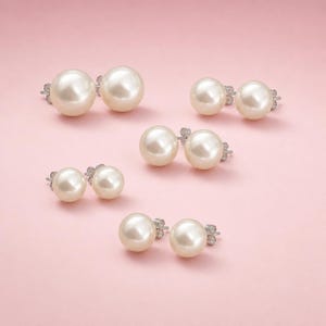 May include: Several pairs of pearl stud earrings are arranged on a pink surface. The earrings feature round, cream-colored pearls with silver-toned posts. The pearls vary slightly in size, offering a classic jewelry option.