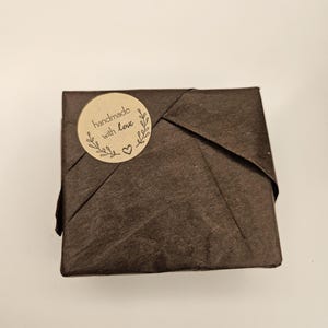 May include: A small, square package wrapped in dark brown paper. A round, tan sticker with the words "handmade with love" and a heart is affixed to the top of the package. The package is likely a gift or a small product.