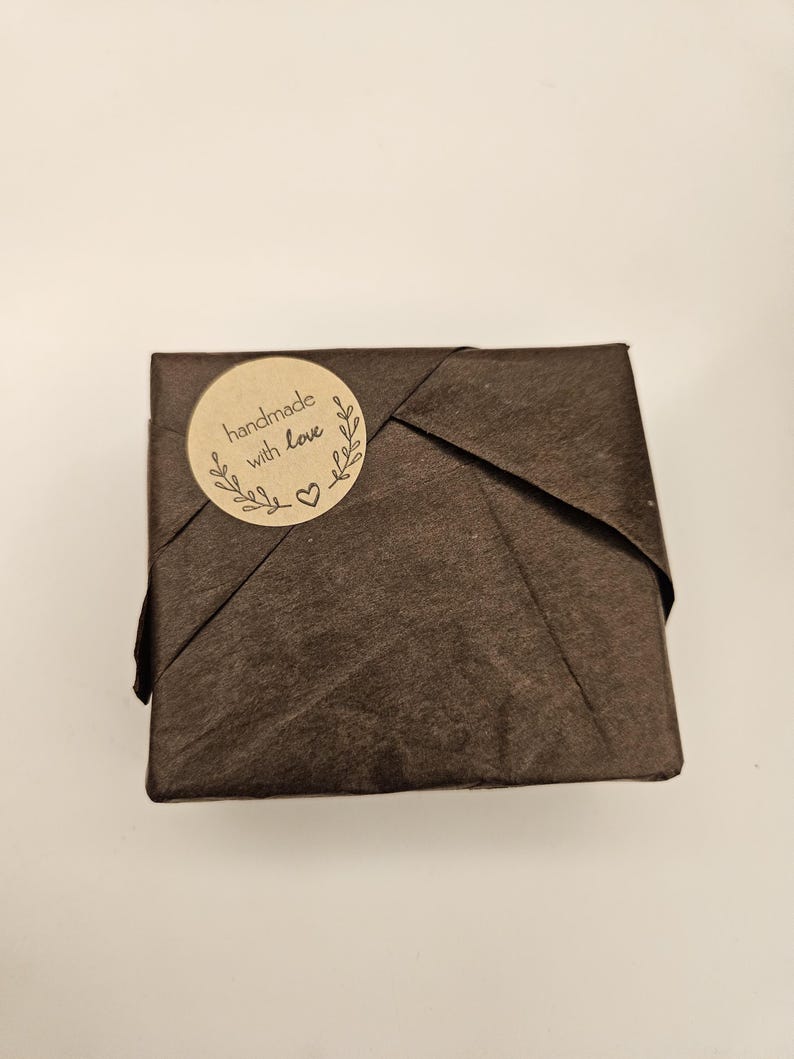 May include: A small, square package wrapped in dark brown paper. A round, tan sticker with the words "handmade with love" and a heart is affixed to the top of the package. The package is likely a gift or a small product.