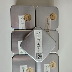 May include: Six small, silver-colored metal tins, some with labels that read "Scent of Time" and "Scent of". Each tin has a small, round sticker with the words "handmade with love". The tins are arranged on a white surface.
