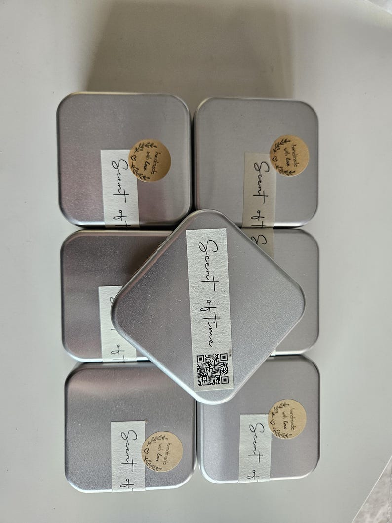 May include: Six small, silver-colored metal tins, some with labels that read "Scent of Time" and "Scent of". Each tin has a small, round sticker with the words "handmade with love". The tins are arranged on a white surface.