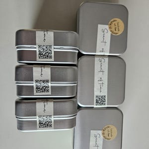 May include: A collection of small, rectangular silver tins, some with the word "Time" printed on a white label and a QR code. Other tins have a sticker that says "Scent of Time".