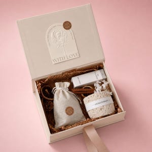 Puede incluir: Caja de regalo crema abierta con un mensaje "With Love" en la tapa. En el interior, una pequeña bolsa con cordón, un tubo blanco, palitos de canela, rodajas de naranja secas y un artículo de aromaterapia redondo.