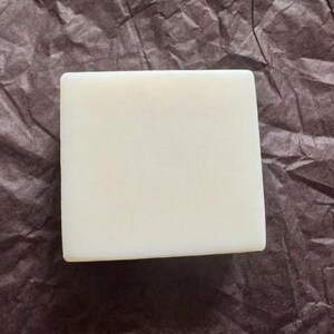 May include: A square, off-white bar of soap. The soap is smooth and has rounded edges. The background is a crumpled, dark brown paper.
