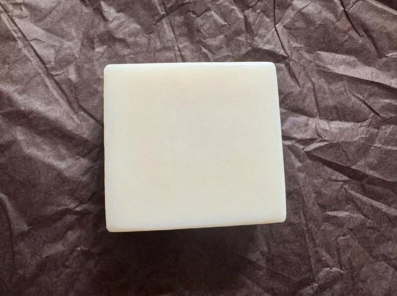 May include: A square, off-white bar of soap. The soap is smooth and has rounded edges. The background is a crumpled, dark brown paper.