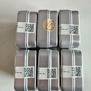 May include: Six rectangular silver metal tins with a vertical stripe down the center. Each tin has a white label with the word "time" and a QR code. One tin has a gold sticker that says "handmade with love".