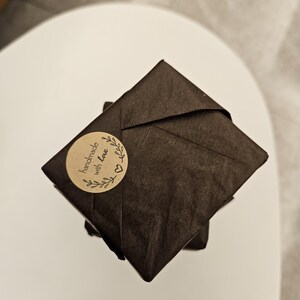 May include: A stack of wrapped packages in dark brown paper. A round sticker with the words "handmade with love" and a heart and leaf design is affixed to the top package. The packages are on a white surface.