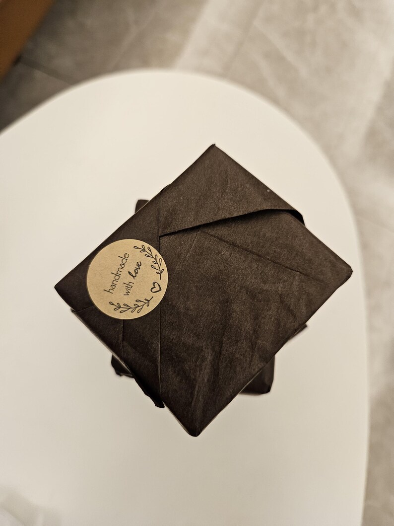 May include: A stack of wrapped packages in dark brown paper. A round sticker with the words "handmade with love" and a heart and leaf design is affixed to the top package. The packages are on a white surface.