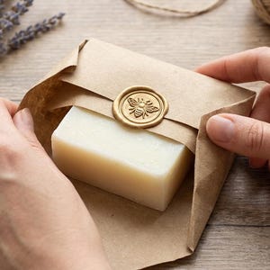 May include: A bar of soap wrapped in brown paper, sealed with a gold bee wax seal. The soap is light in color and the packaging is held by two hands. Dried lavender and twine are in the background.