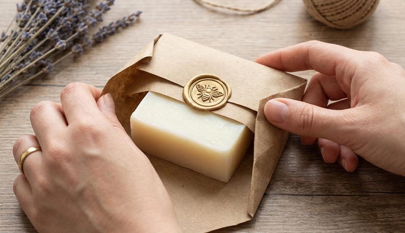 May include: A bar of soap wrapped in brown paper, sealed with a gold bee wax seal. The soap is light in color and the packaging is held by two hands. Dried lavender and twine are in the background.