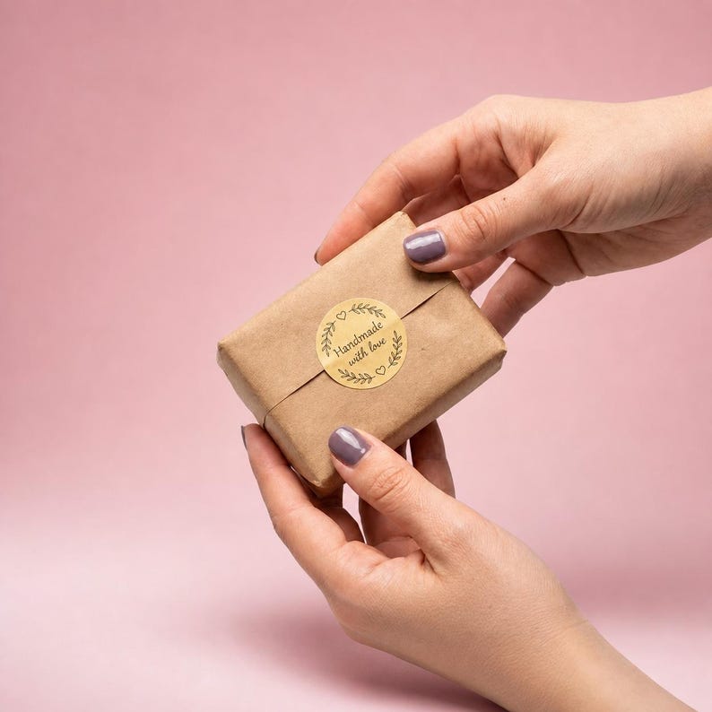 May include: A small, rectangular package wrapped in brown paper, held by two hands against a pink background. A gold sticker with the words "Handmade with love" and a wreath design is affixed to the package.