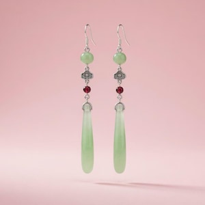 Op de afbeelding: Een paar oorhangers met lichtgroene jade druppelvormen. De oorbellen hebben zilverkleurige haken, bloemaccenten en rode kralen. De achtergrond is effen roze.