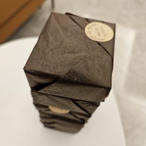 May include: A stack of three dark brown, wrapped packages. Each package is sealed with a small, round sticker that reads "handmade with love" in cursive script. The packages are stacked on a white surface.