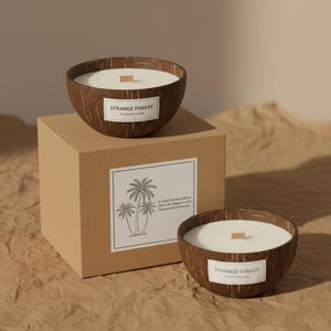 Puede incluir: Dos velas perfumadas en cáscaras de coco con cera blanca y mechas de madera, etiquetadas como "Strange Forest Scented Candle". Una vela está sobre una caja marrón con un gráfico de palmera y texto.