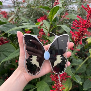 Könnte beinhalten: Ein Buntglas-Schmetterlings-Sonnenfänger mit schwarzen und weißen Flügeln und einem blauen Glas-Mittelteil. Der Sonnenfänger hängt an einer silbernen Kette.